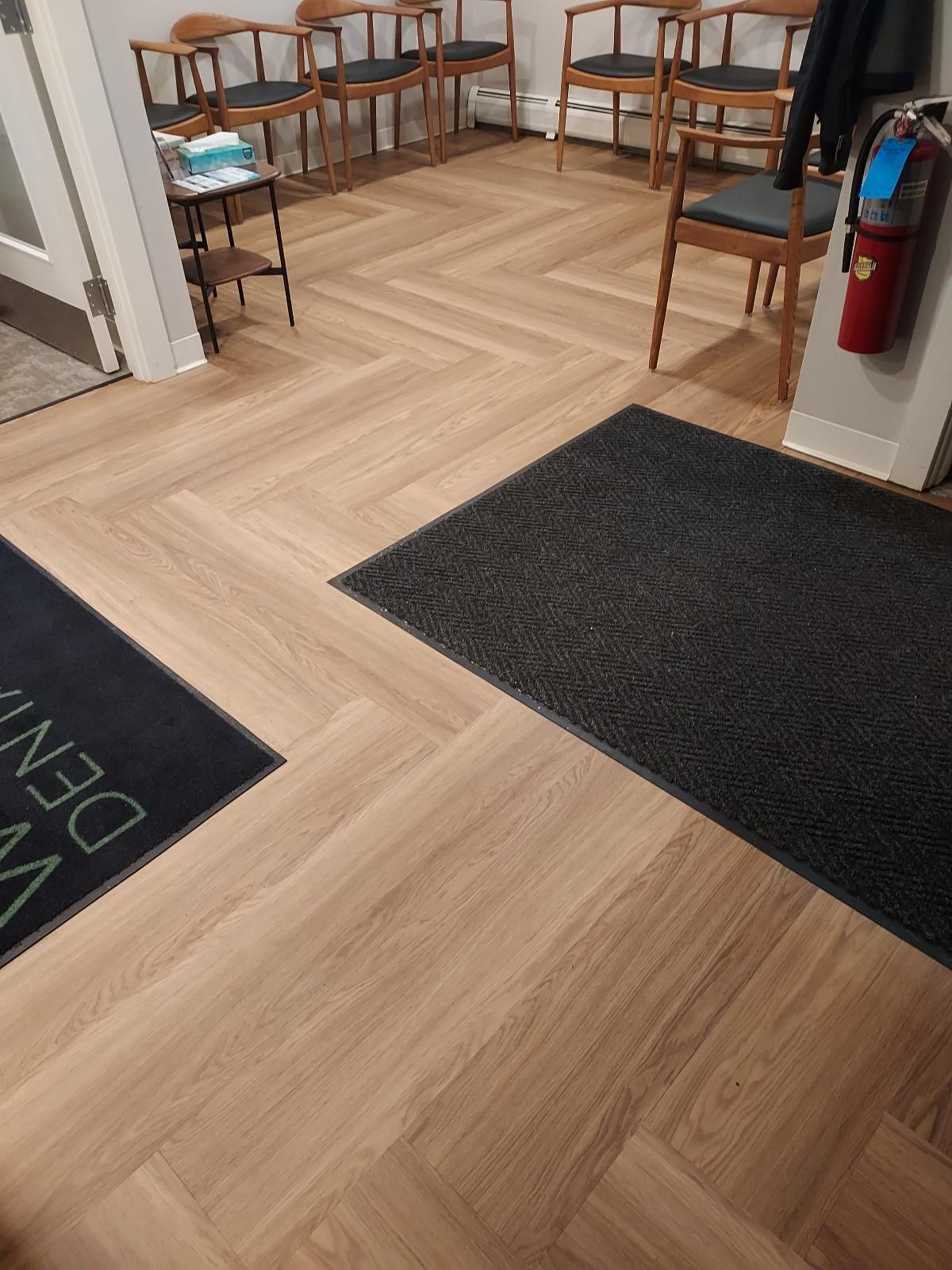 A waiting room with light oak-look flooring in a herringbone pattern, featuring several wooden chairs, dark entrance mats, and a wall-mounted fire extinguisher.