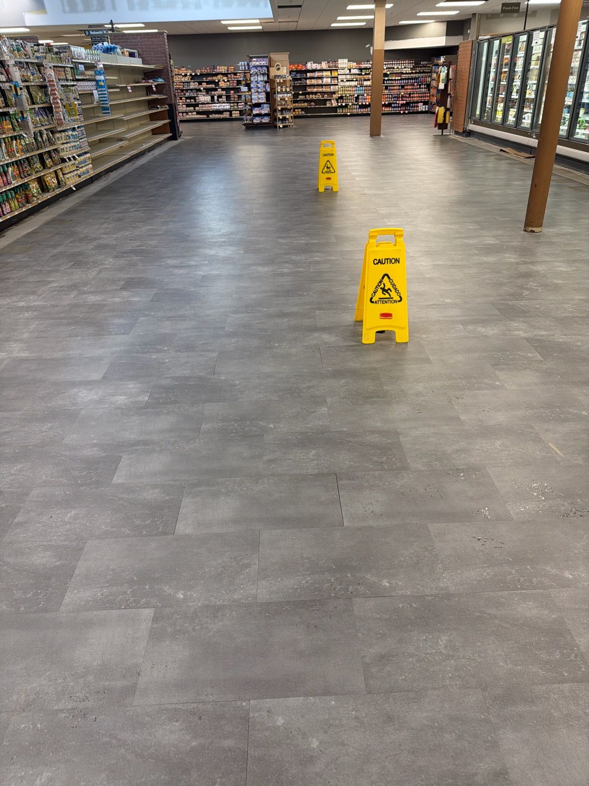 A grocery store aisle with grey stone-look vinyl flooring and two yellow caution signs positioned between empty and stocked shelves.