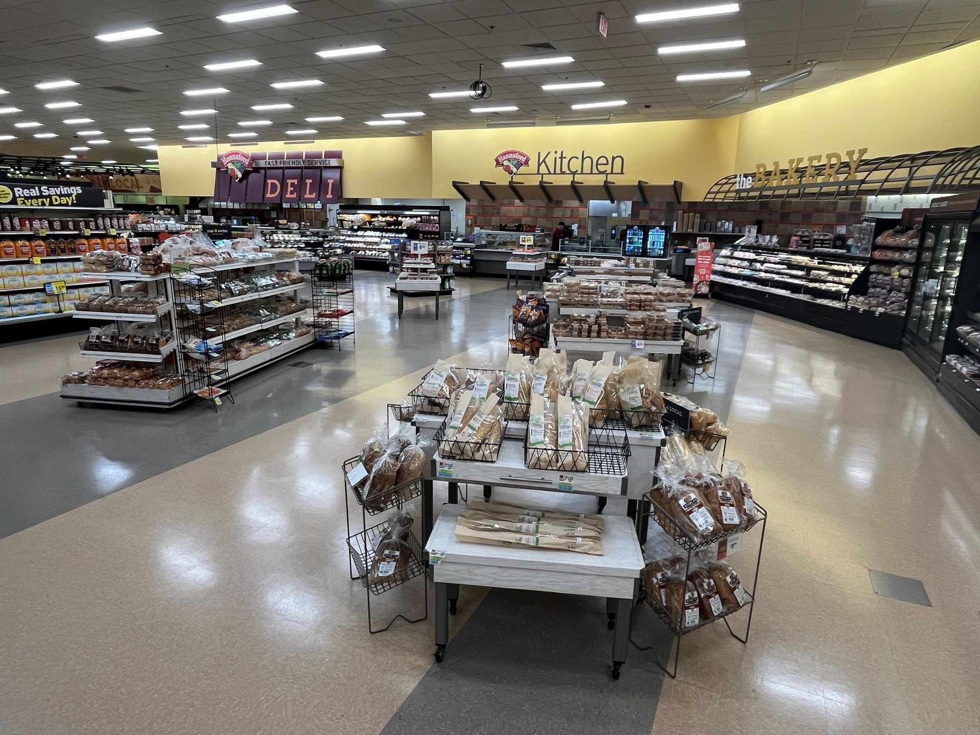 Grocery store aisle with shelves of packaged goods and a central bakery display under bright ceiling lights