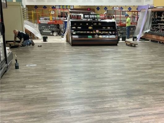 Store interior with wood-look flooring, shelves, and a worker stocking items near the left wall