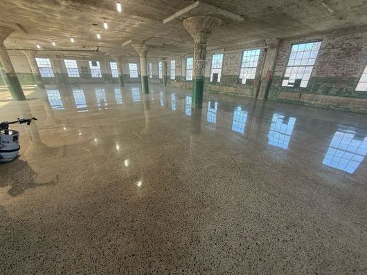 Large empty industrial room with polished concrete floors, brick walls, and tall windows reflecting light