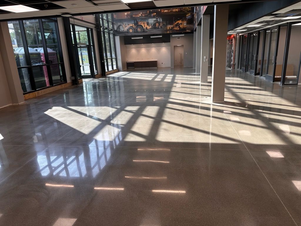 A large, empty commercial indoor space with a highly polished concrete floor reflecting bright sunlight from large windows.