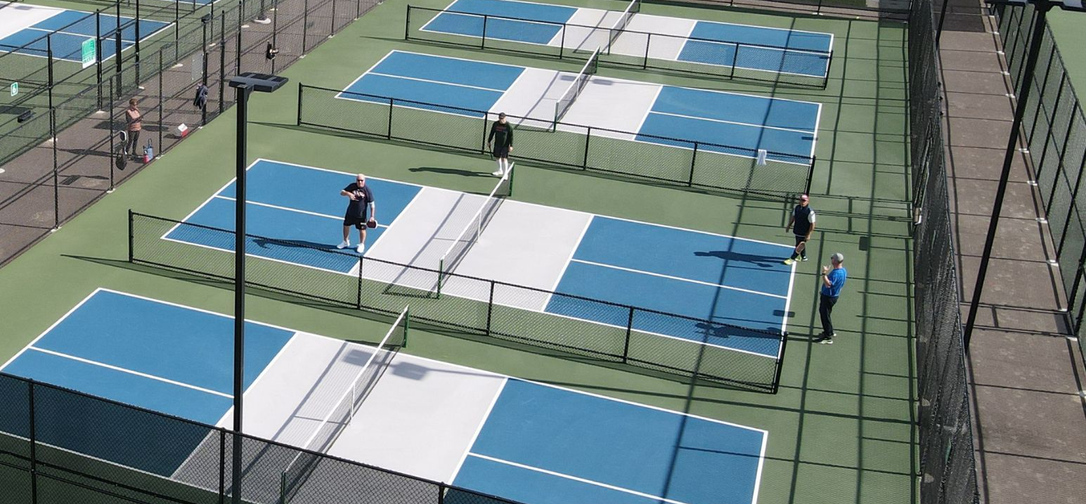 Pickleball players on blue and white courts. Green grass surrounds the courts.