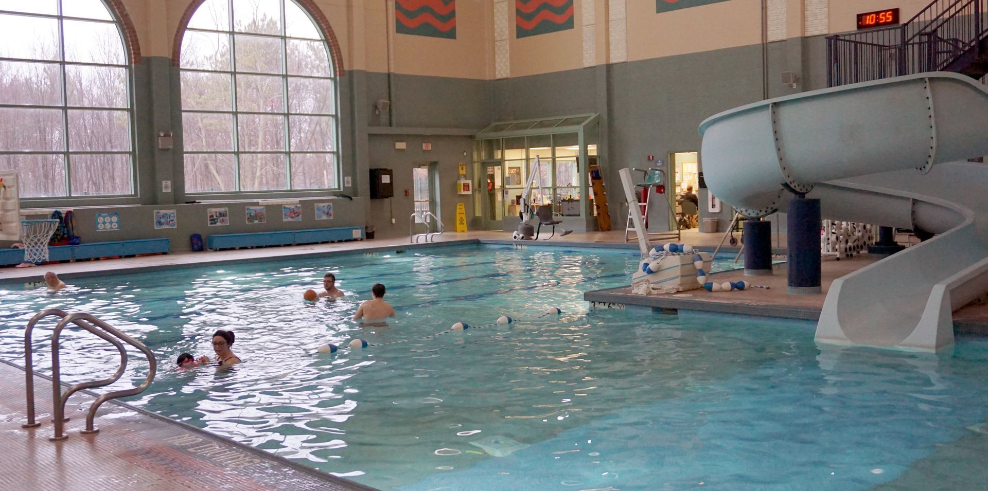 Indoor swimming pool with people in the water, a slide, and large arched windows.
