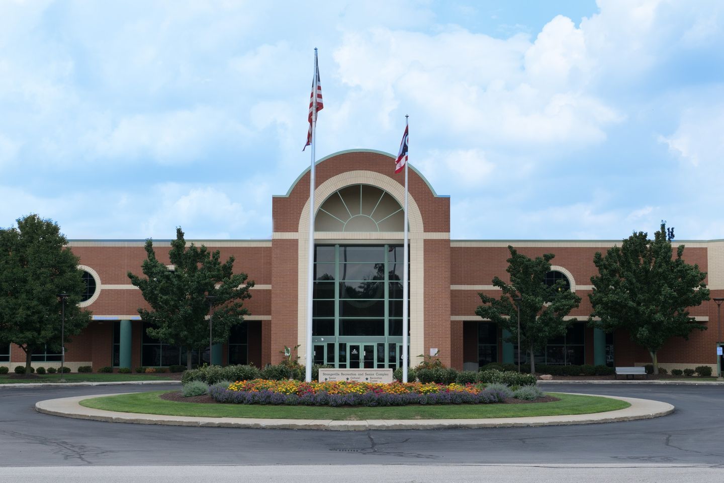 Brick building with arched entrance and flags, circular driveway with flowers.