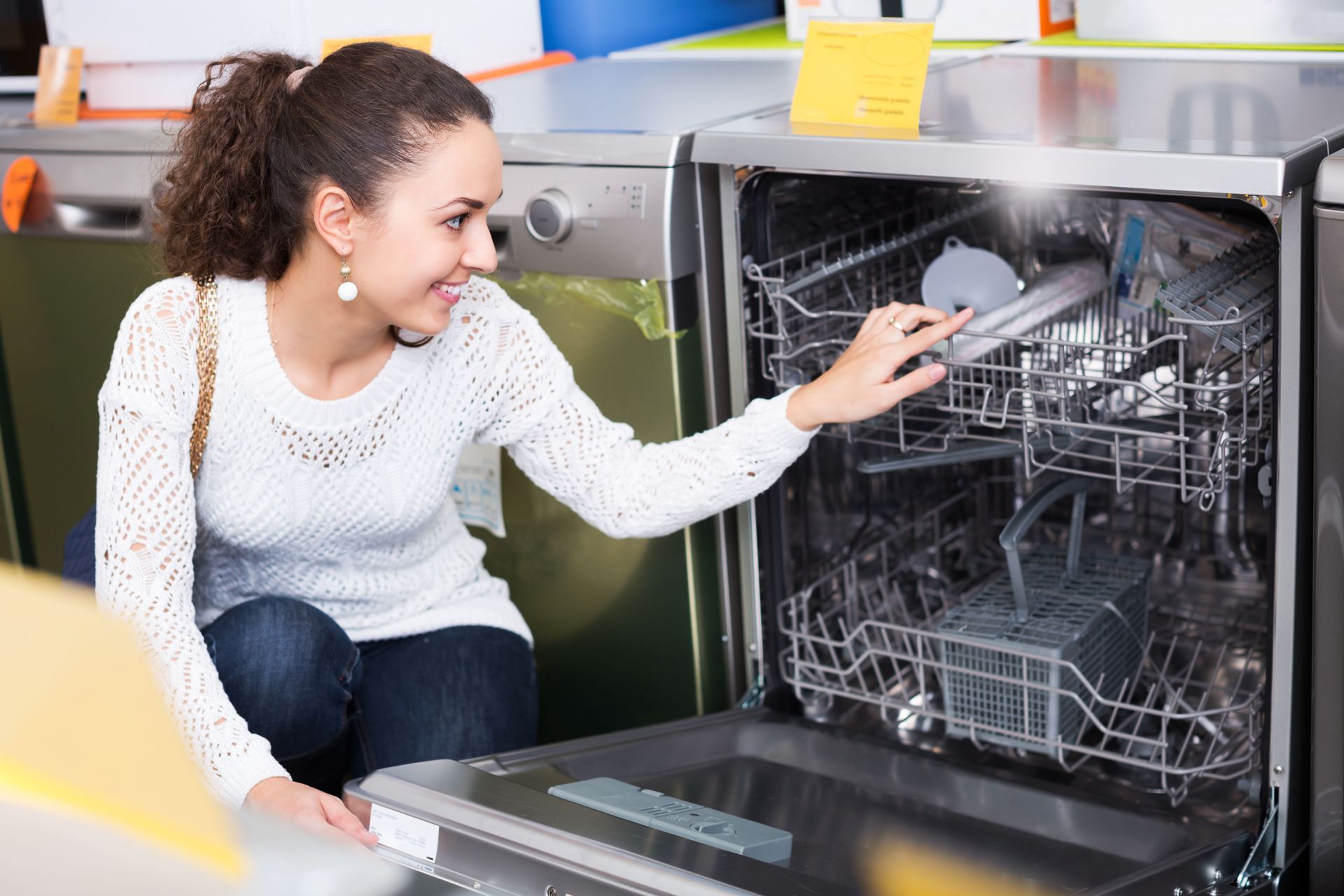 Woman examining a dishwasher in an appliance store; smiling, reaching inside.