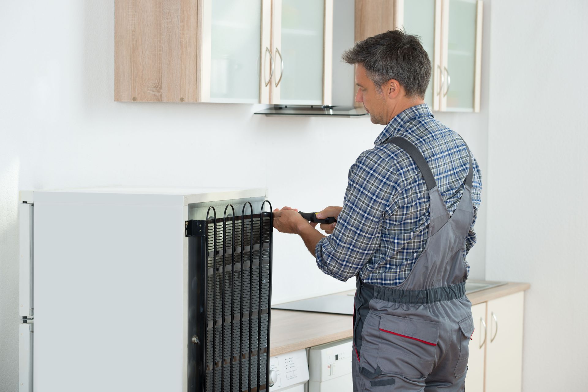 Man in overalls repairing a refrigerator in a kitchen.
