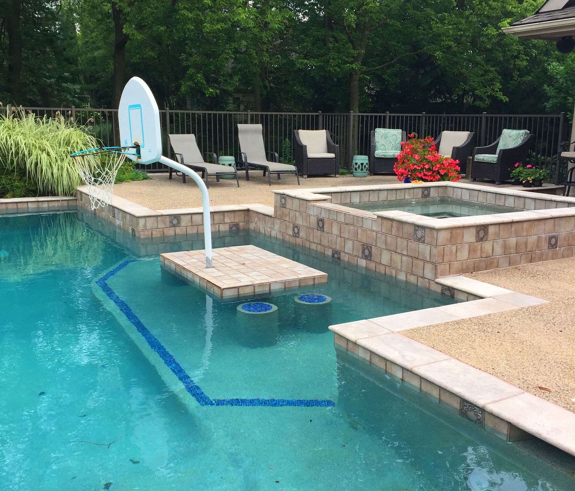 Backyard pool with basketball hoop, hot tub, and lounge chairs. Turquoise water and tan stonework.