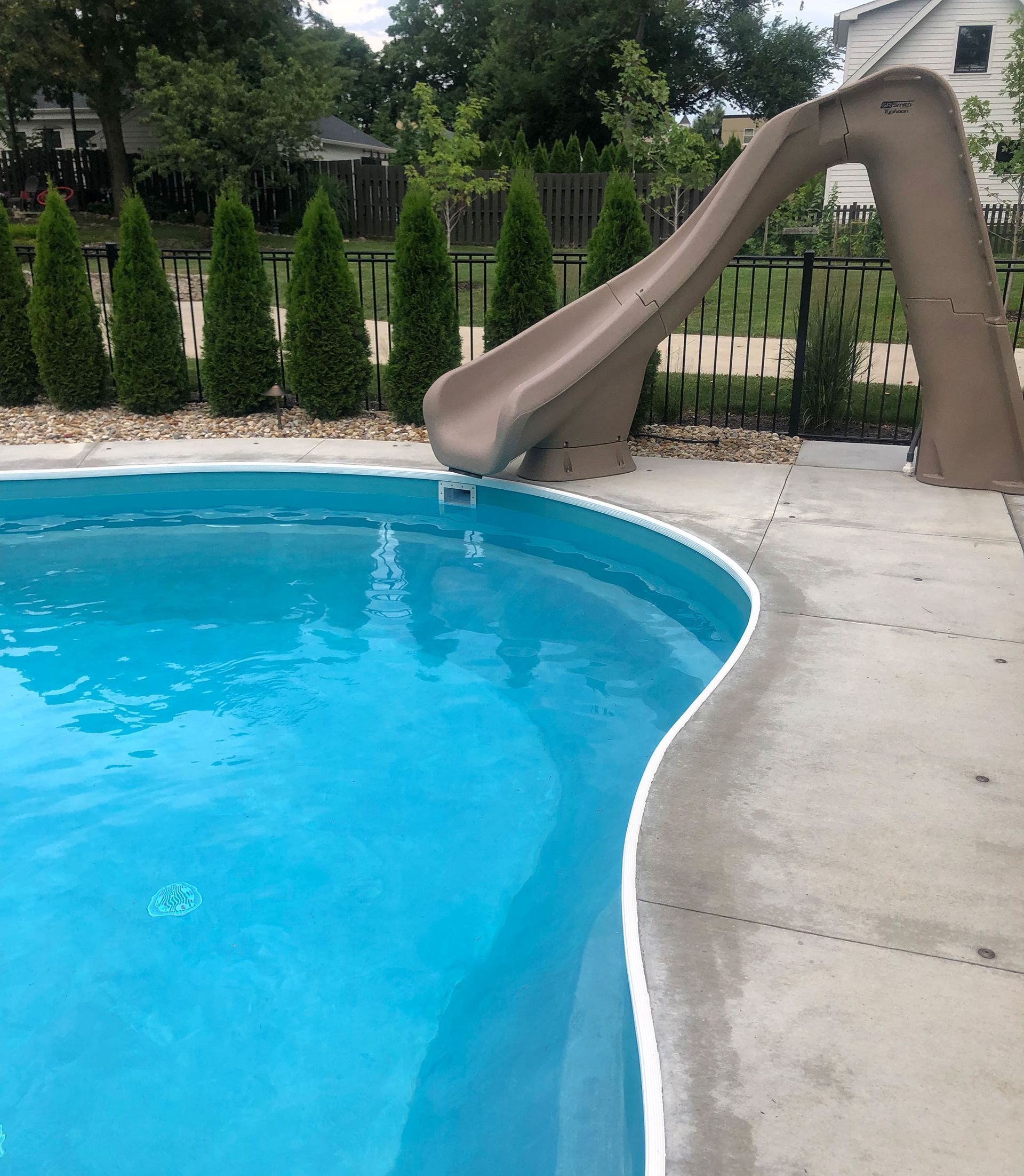 Swimming pool with brown slide; blue water, concrete deck, and green trees.