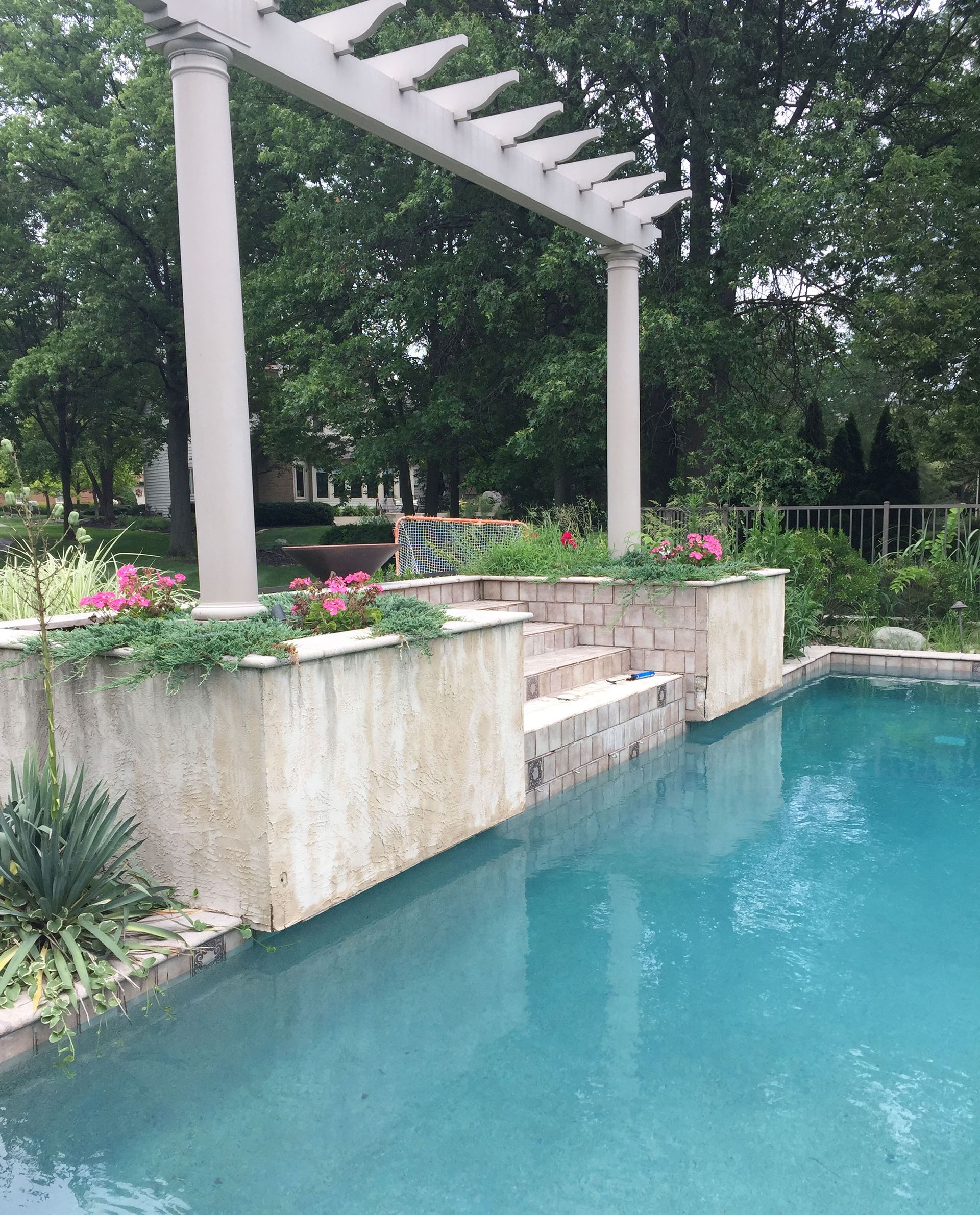 Poolside pergola with steps, planters, and teal water. Green trees in the background.