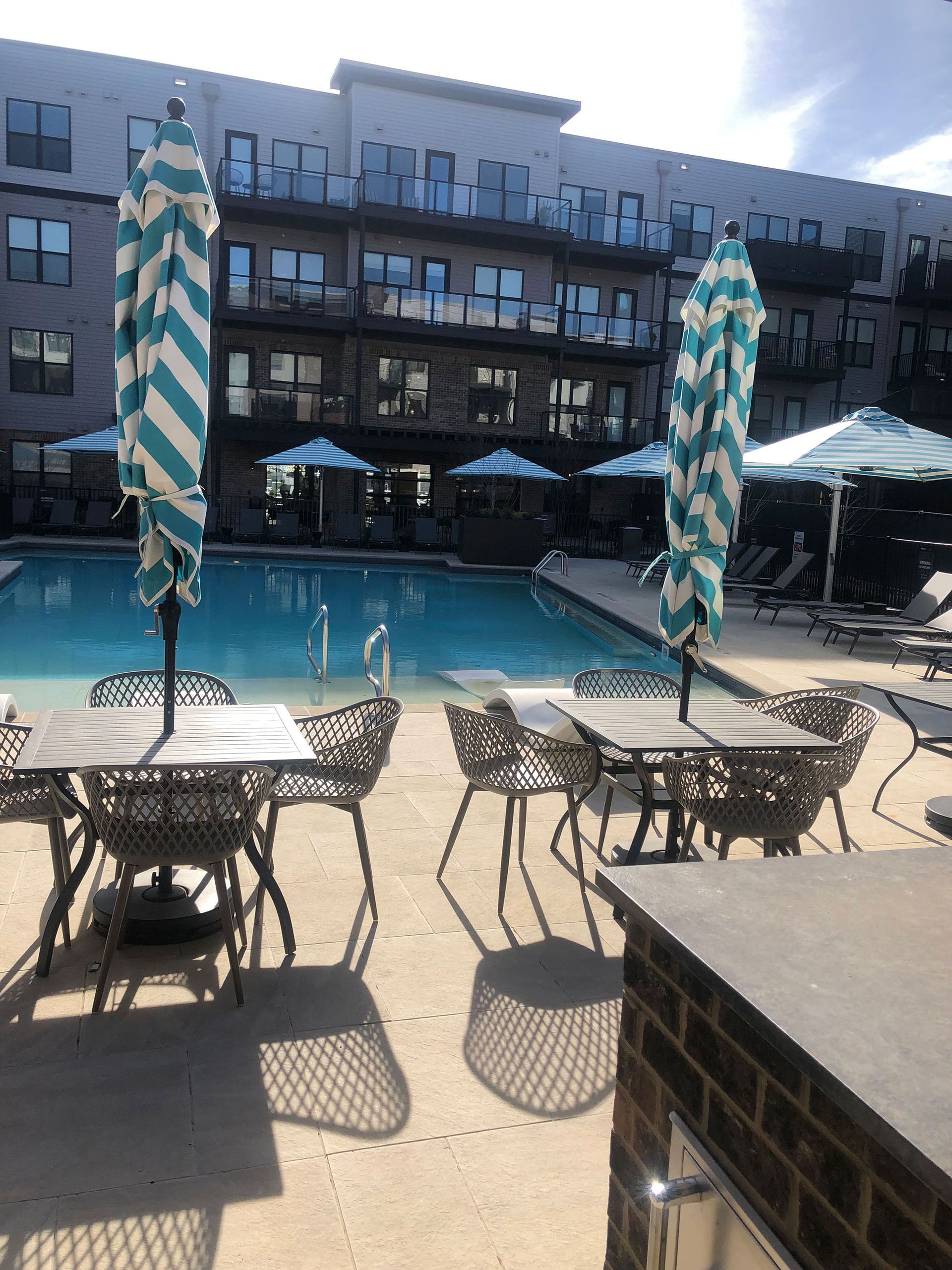 Poolside patio with tables, chairs, and striped umbrellas. Modern building in background. Sunny day.