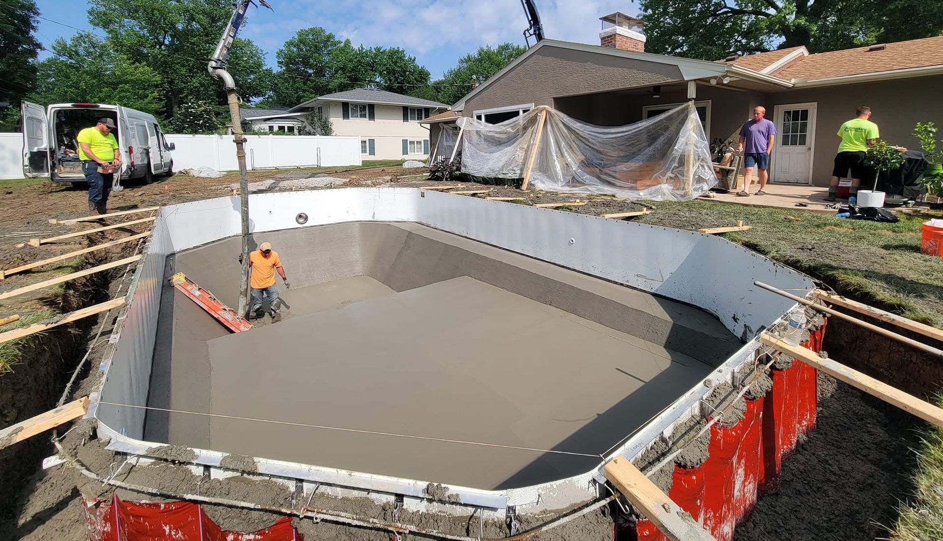 Construction of a swimming pool: workers pouring concrete, white pool walls, and a house in the background.