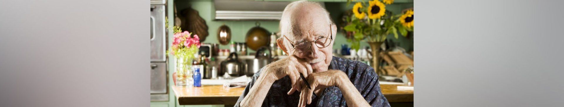 An older man is sitting in a kitchen with his hands folded.