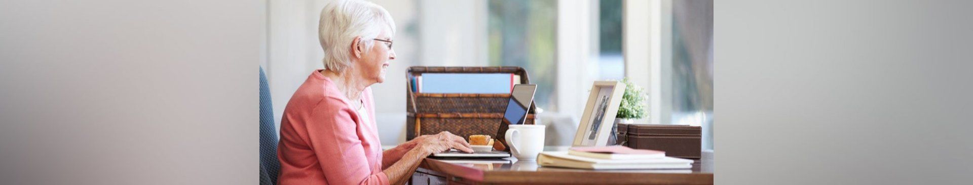 An elderly woman is sitting at a desk using a laptop computer.
