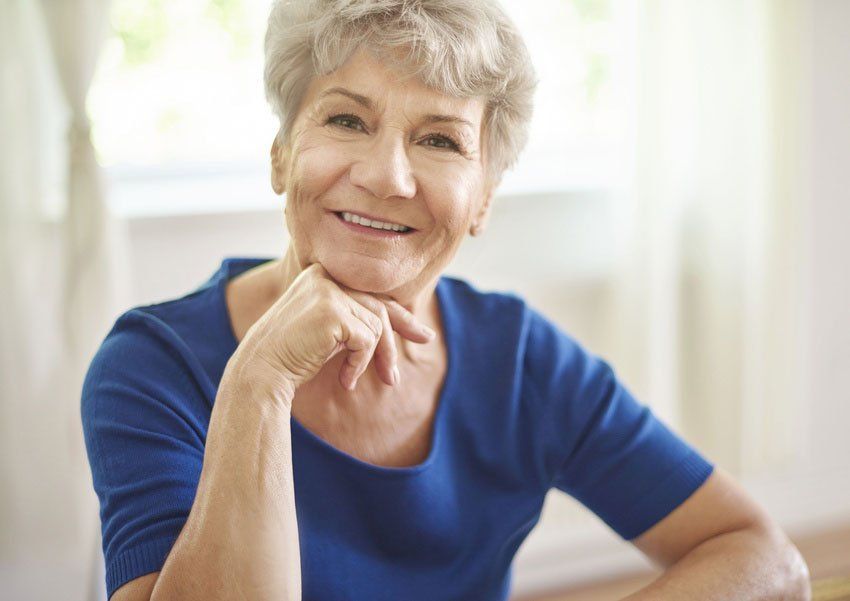 An elderly woman in a blue shirt is smiling with her hand on her chin.