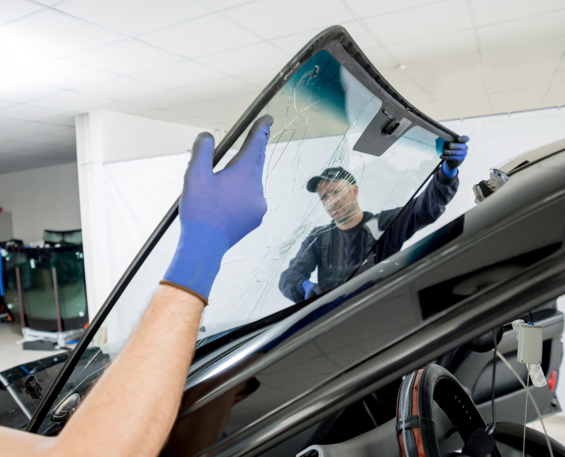 A person replacing a car windshield inside a well-lit workshop. Two hands wearing blue gloves handle the glass.