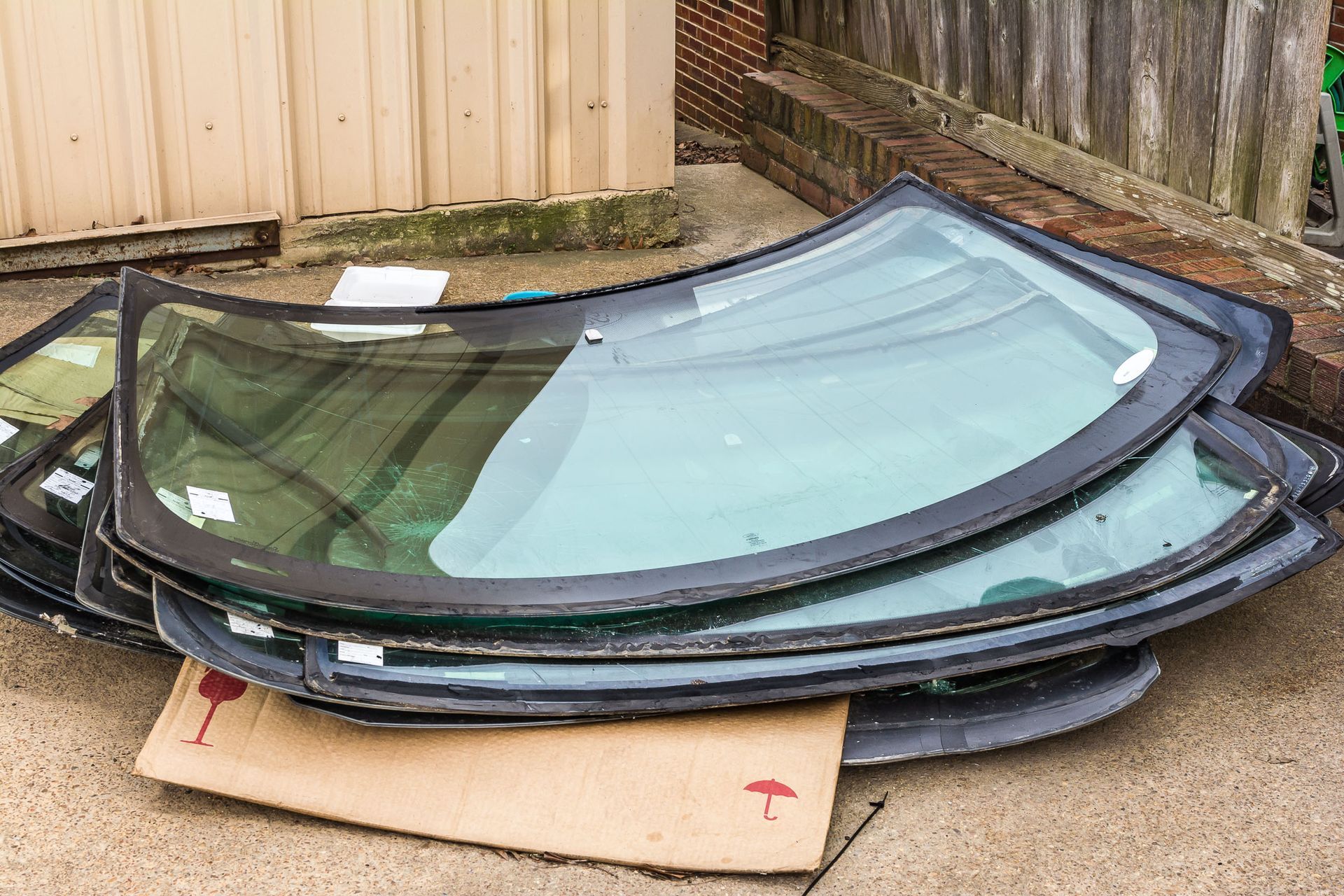 Pile of used automotive windshields on cardboard next to a weathered wall and wooden fence.