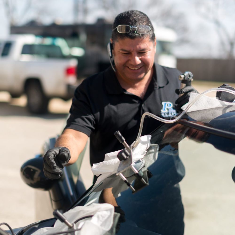 A person in a black shirt working on a car windshield, smiling.  Outdoor setting with a truck visible.