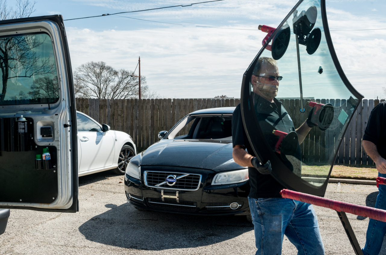 A man carrying a car windshield, next to a black Volvo and a white car in a sunny outdoor setting.