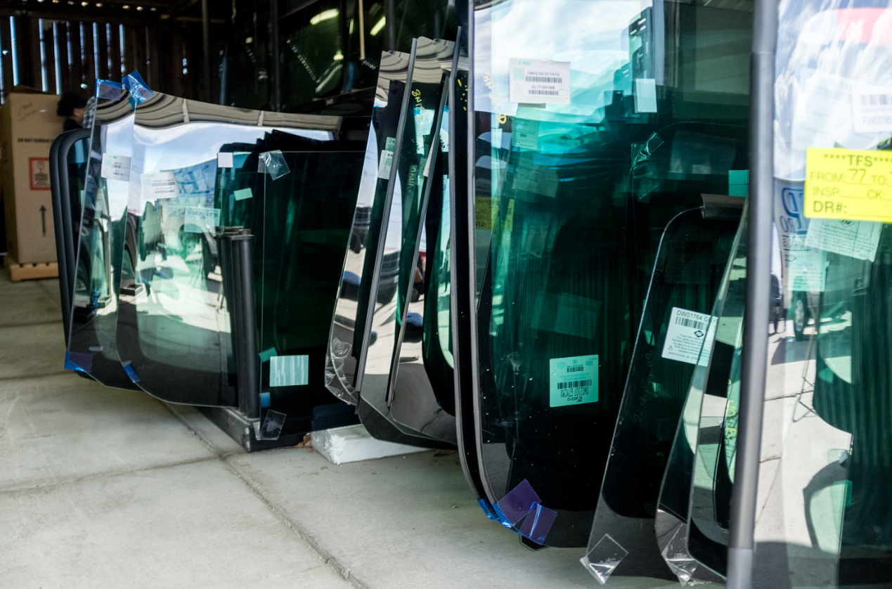 Windshields stacked in a warehouse, reflecting a cityscape. Green tint, labels attached.