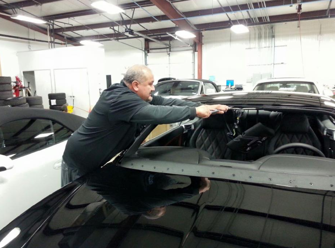 Man working on a car in a shop, installing part of the roof. Black car with interior visible.
