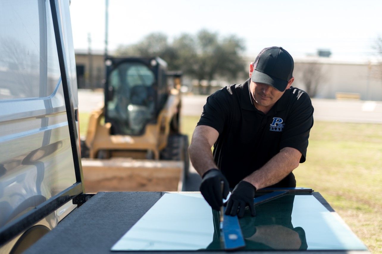 Man in black shirt and cap working on a window pane on a van, with a yellow construction vehicle in the background.