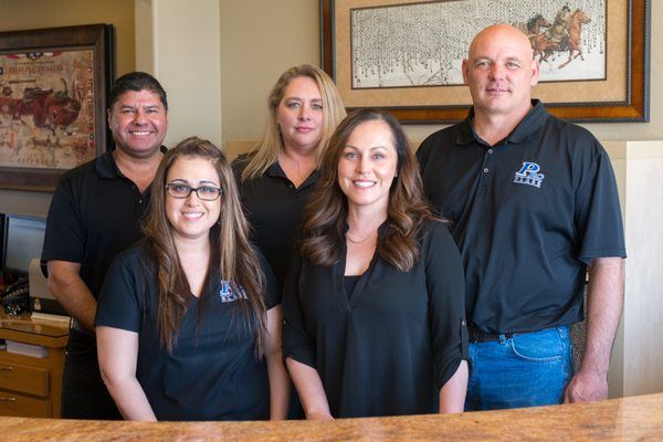 Five people in black shirts smiling at the camera, standing behind a wooden counter. Artwork in the background.