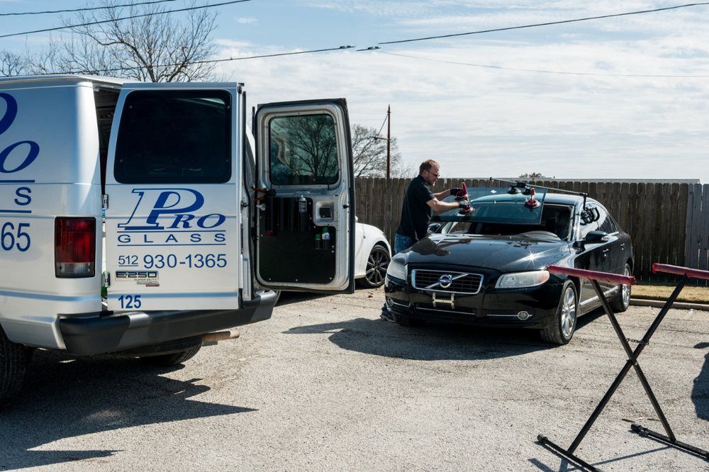 A technician replacing a windshield on a black Volvo, parked beside a Pro Glass van.