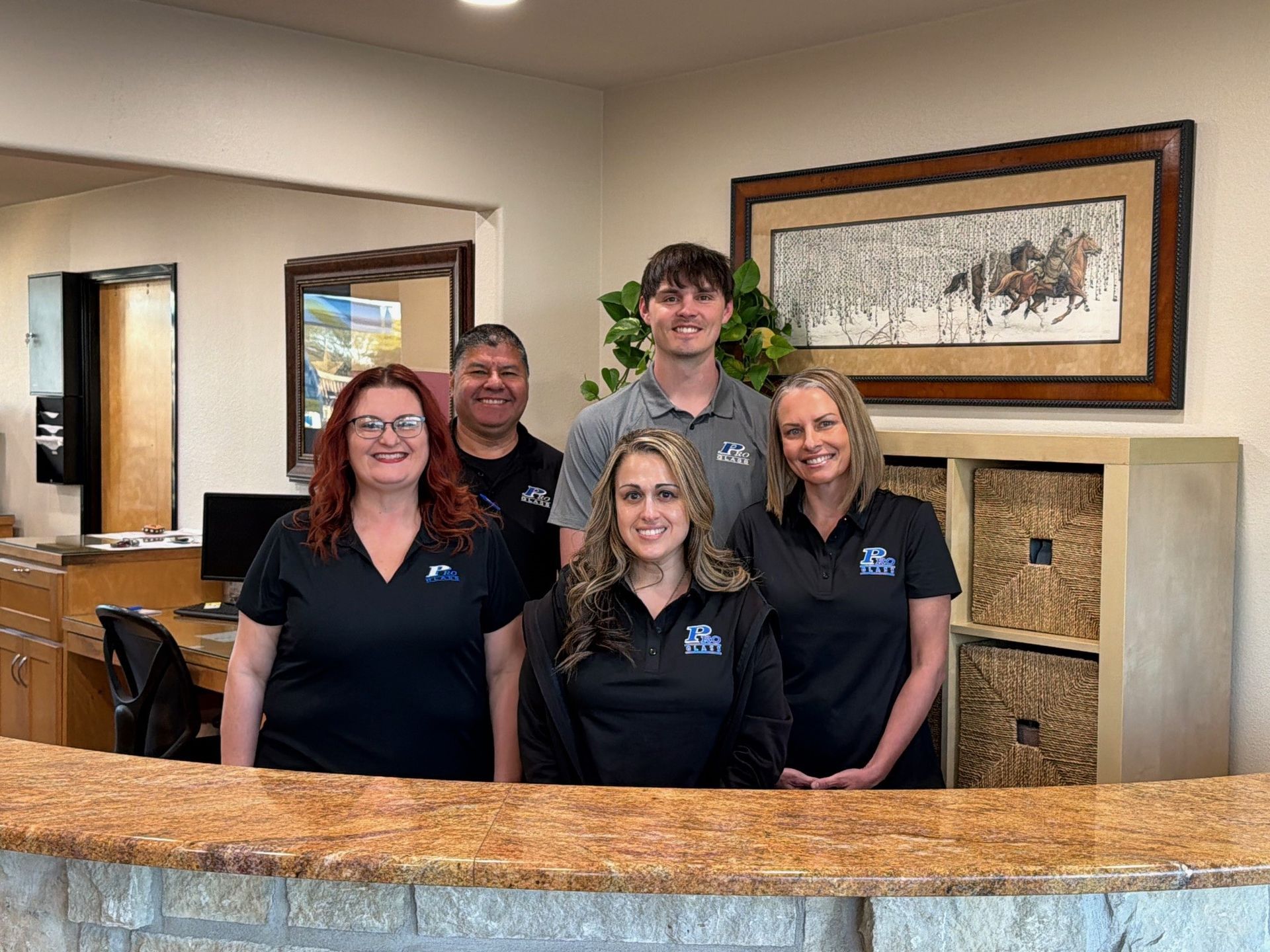 Five staff members in black shirts smiling behind a front desk in an office with framed artwork on the wall.