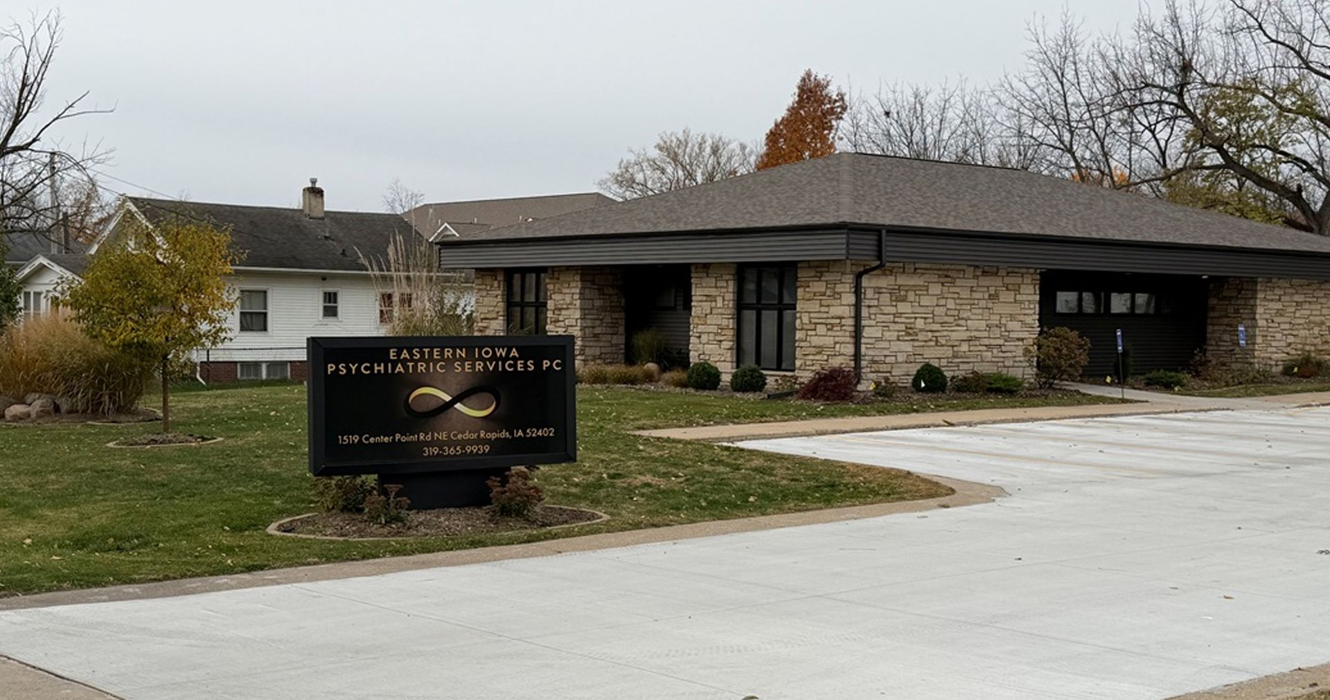 Sign for a business with a light brick building, next to a house, on a cloudy day.