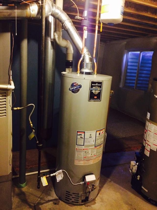 Water heater in a basement. Cylindrical, silver tank with exhaust pipe. Dark setting.
