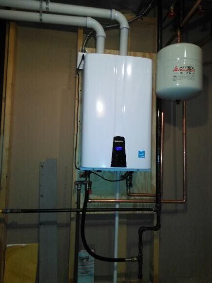 White tankless water heater mounted on a wooden wall, with connected pipes and an expansion tank.