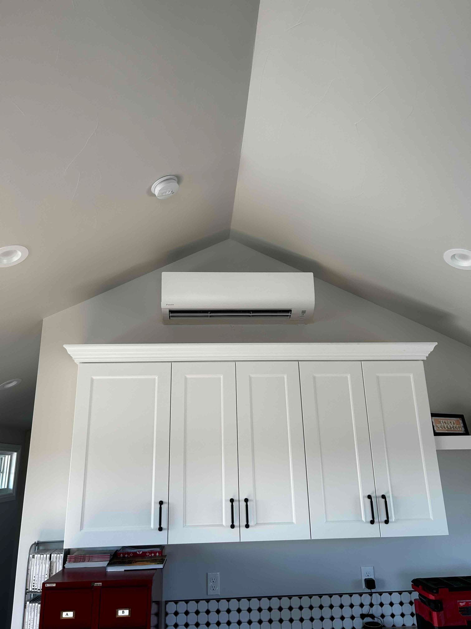 White air conditioning unit above white kitchen cabinets. Grey ceiling.