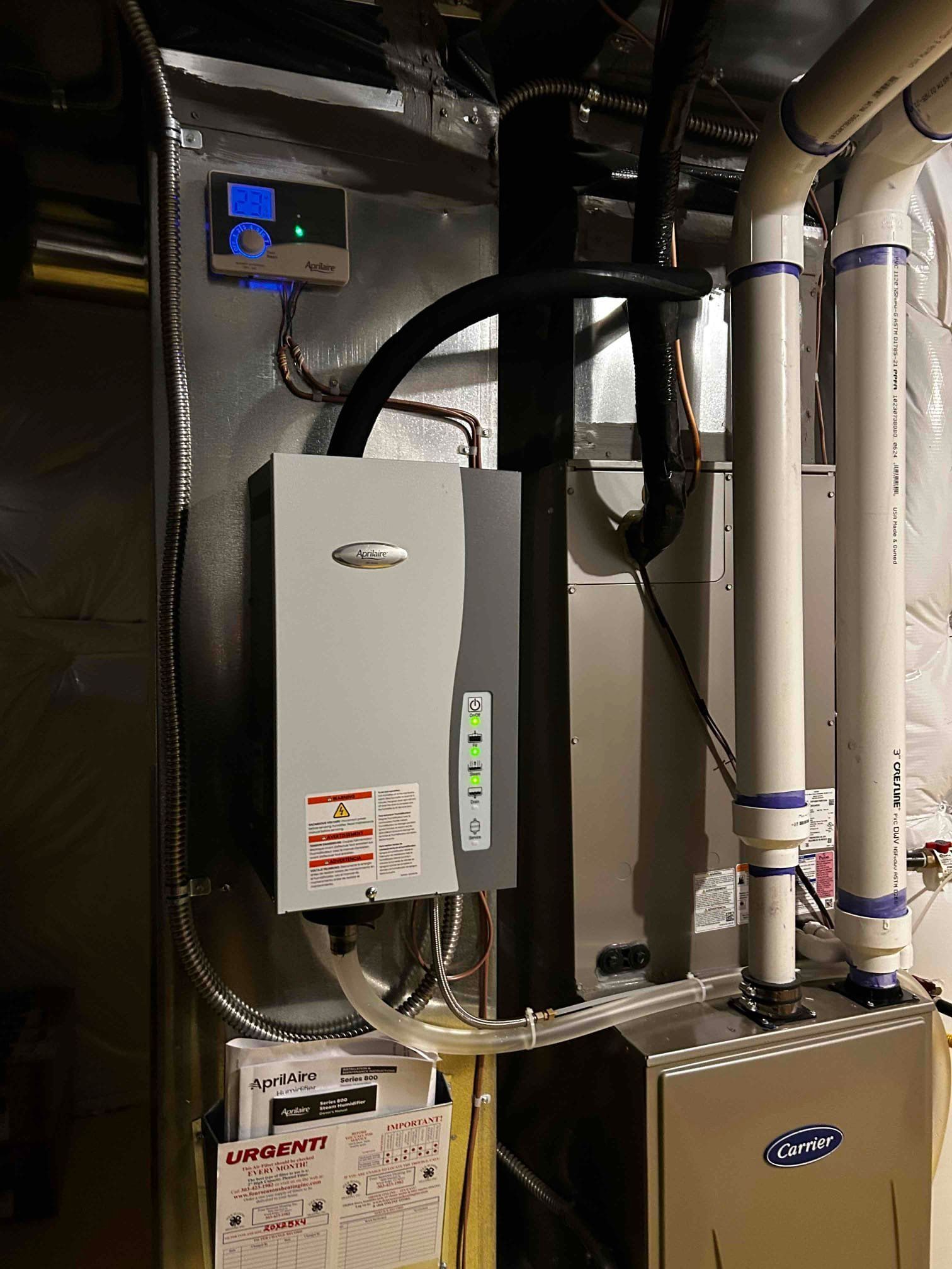 Humidifier unit installed on a furnace in a utility room. Metal ducts and pipes are visible.