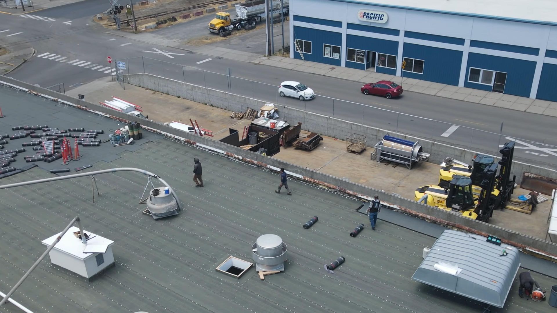 Rooftop scene with people and equipment. Cars and a building are in the background. Gray and blue tones are present.