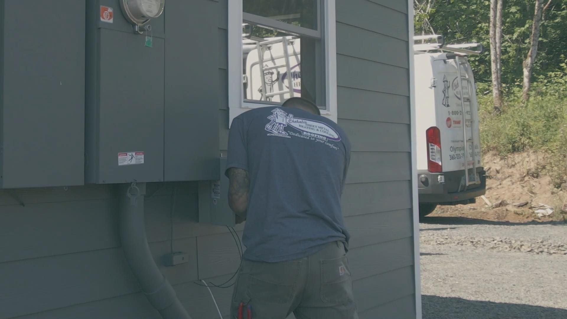 An electrician works on a power box attached to a gray building. A white work van is parked nearby.