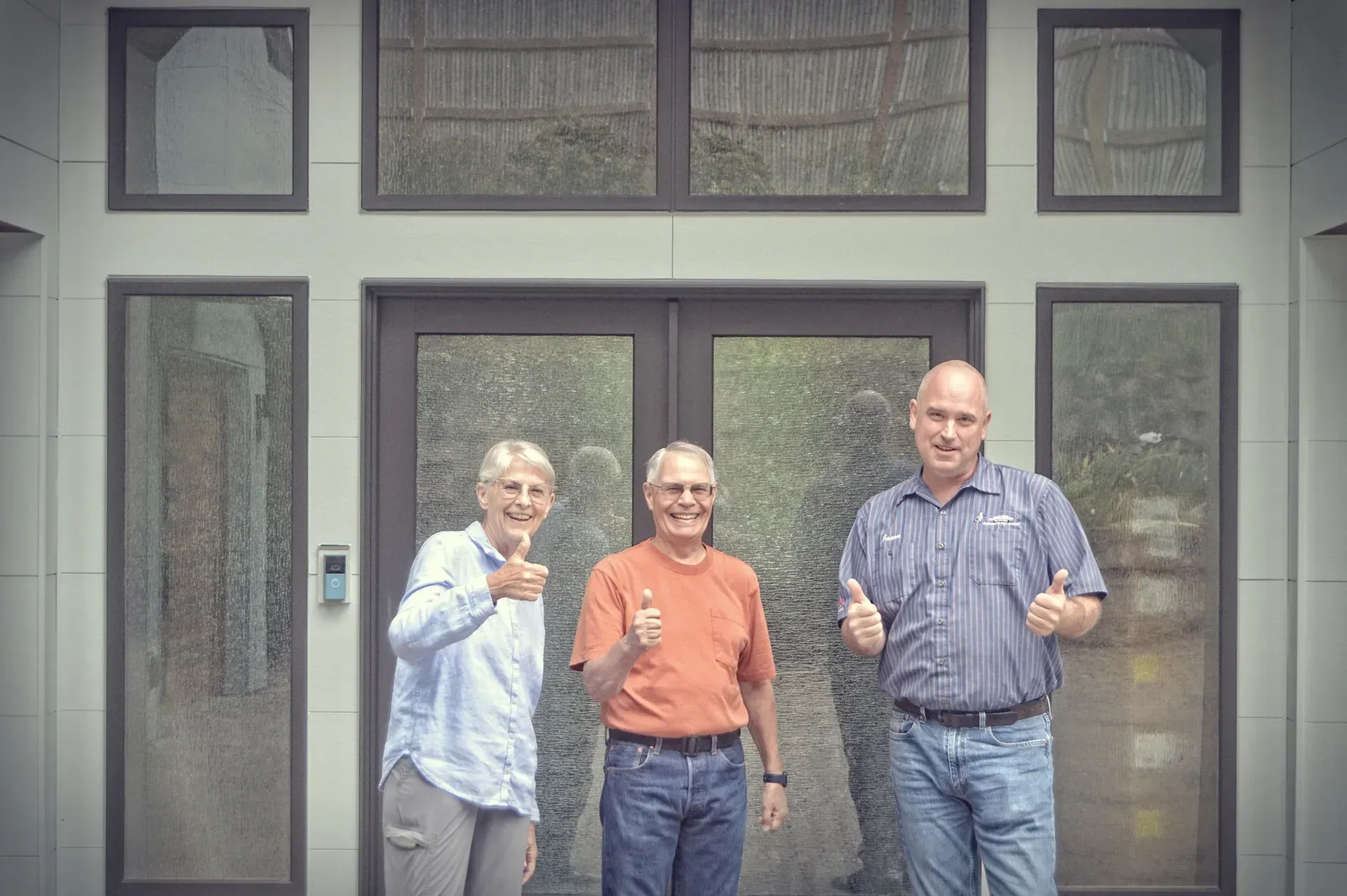 Three people giving thumbs-up in front of a building with glass doors and windows; they appear happy.