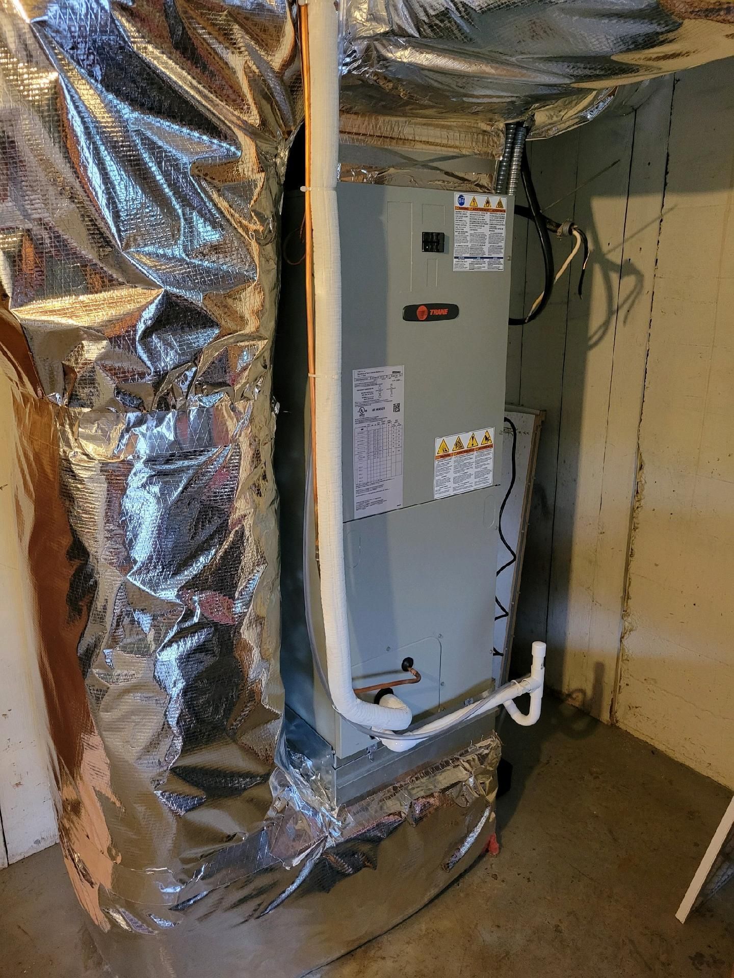 HVAC unit in a basement. Ductwork wrapped in silver insulation. White pipes. Concrete walls and floor.
