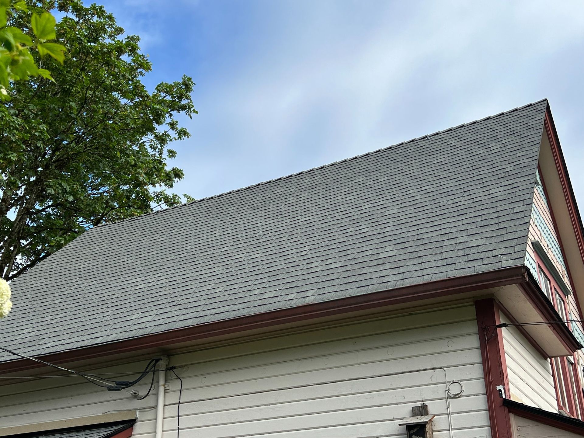 Gray shingled roof of a house against a cloudy sky. Light-colored siding with red trim.