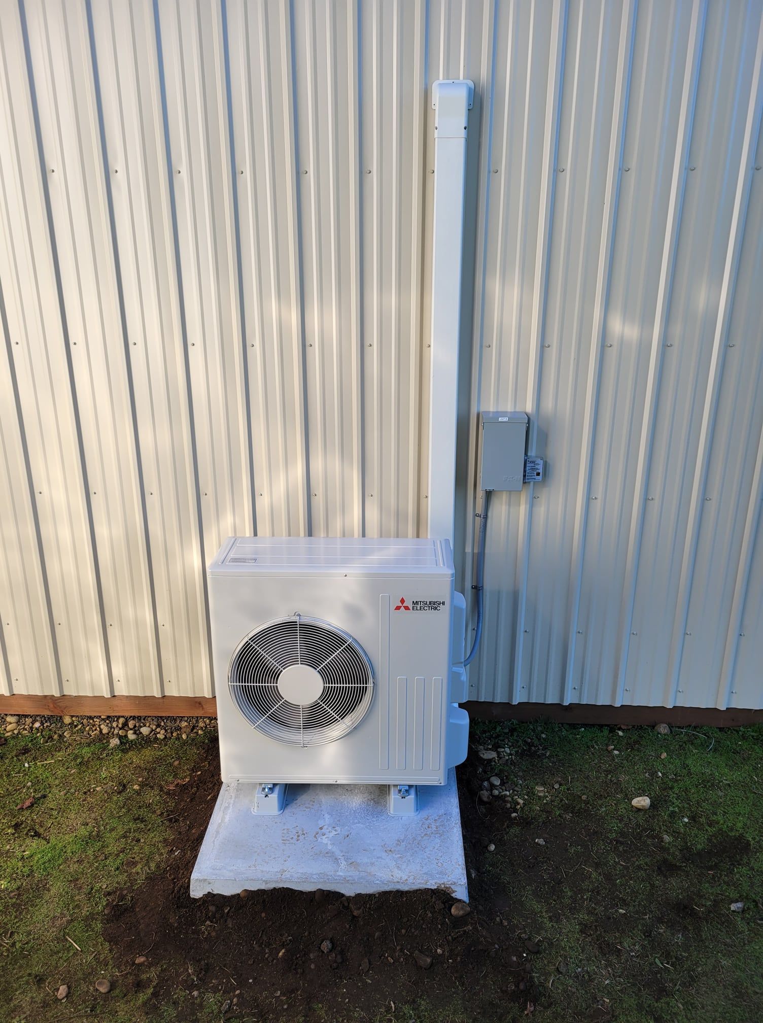 White outdoor air conditioning unit mounted on a concrete slab, against a white corrugated metal wall.