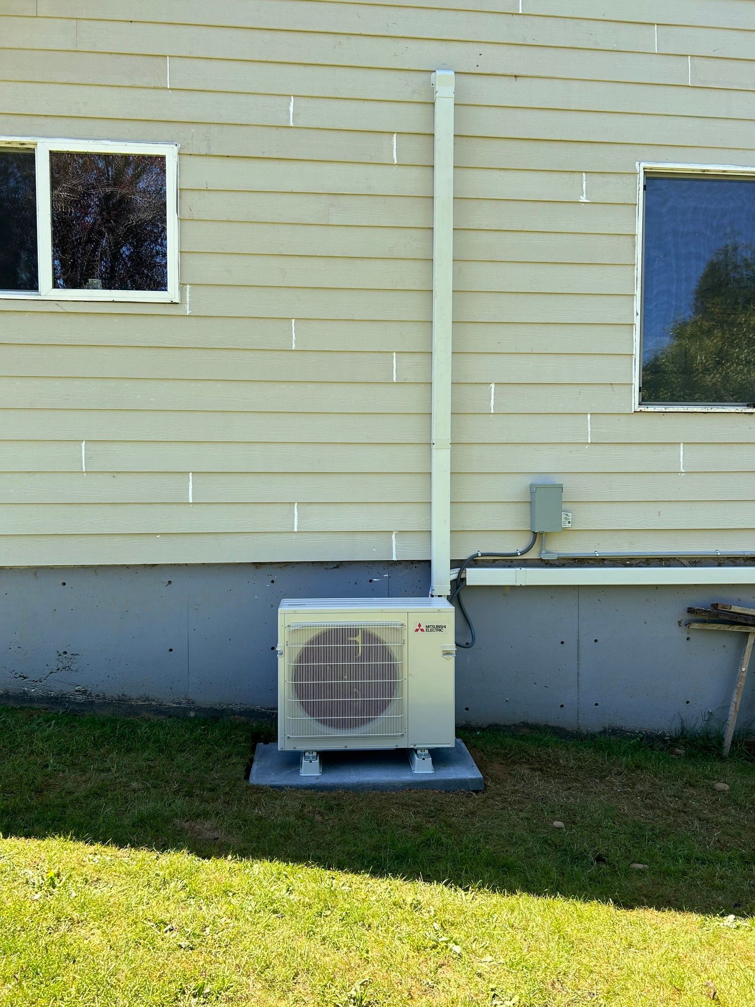 An outdoor air conditioning unit on a concrete pad near a house. White siding, white conduit.