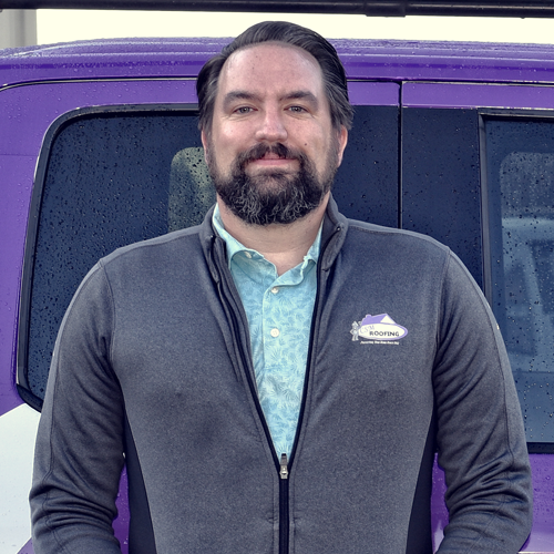 Man in a grey jacket and blue shirt stands in front of a purple vehicle.