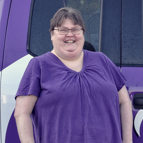 Woman wearing glasses and purple shirt smiles, standing beside a purple vehicle.