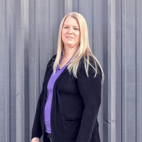 Woman with blonde hair wearing a purple shirt and black cardigan stands in front of a gray wall.