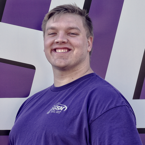 Man in purple shirt smiling in front of a purple and white backdrop.