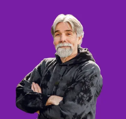 Man with grey beard and hair, arms crossed, wearing a black tie-dye hoodie, purple background.