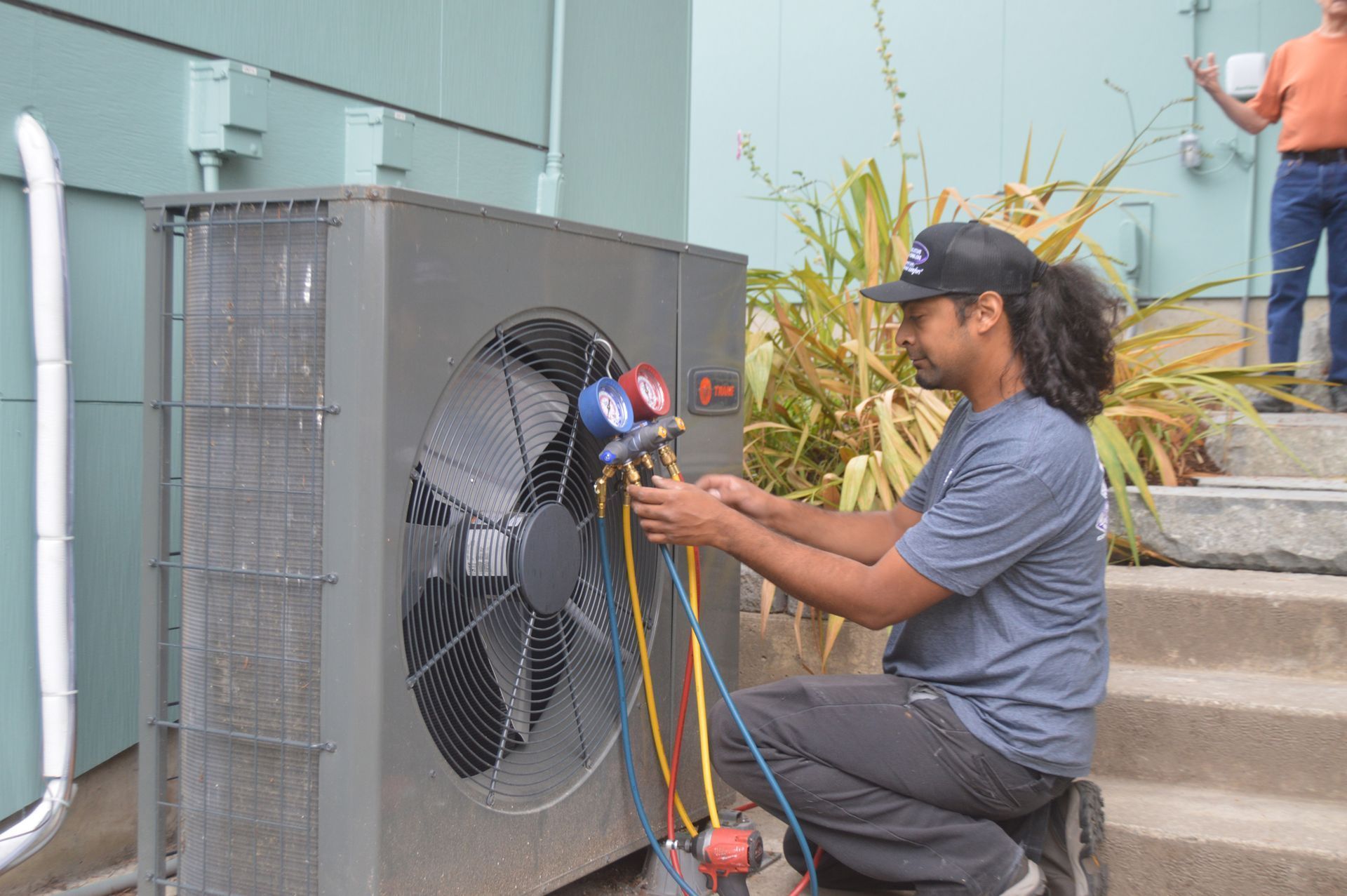 HVAC technician connects gauges to an outdoor heat pump unit. Yellow, blue, and red hoses are visible.