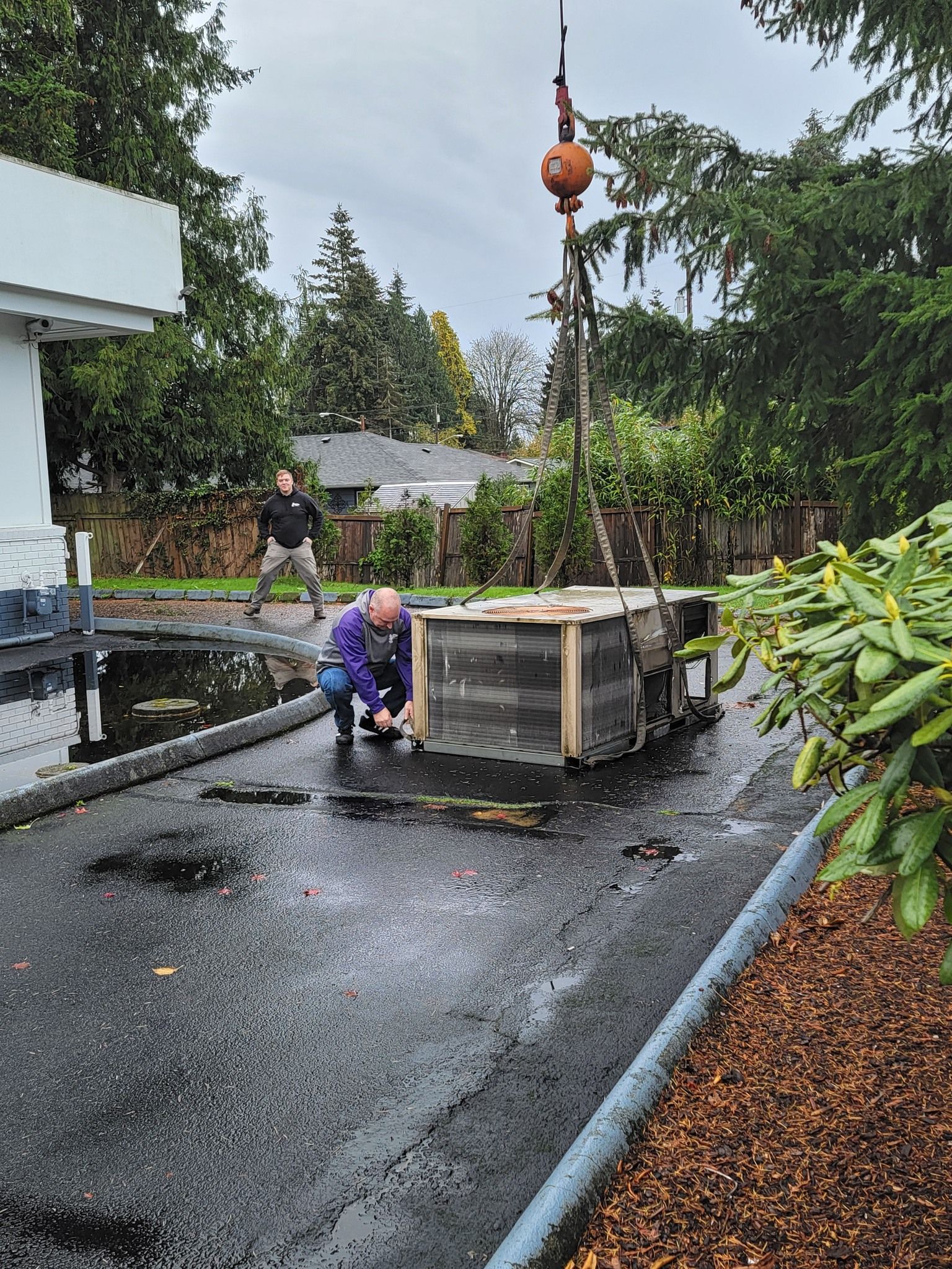Two people maneuvering a large crate on a wet driveway, suspended by a crane. Overcast sky.
