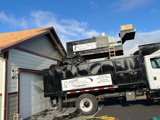 Black dump truck next to a garage, filled with debris. Blue sky overhead.
