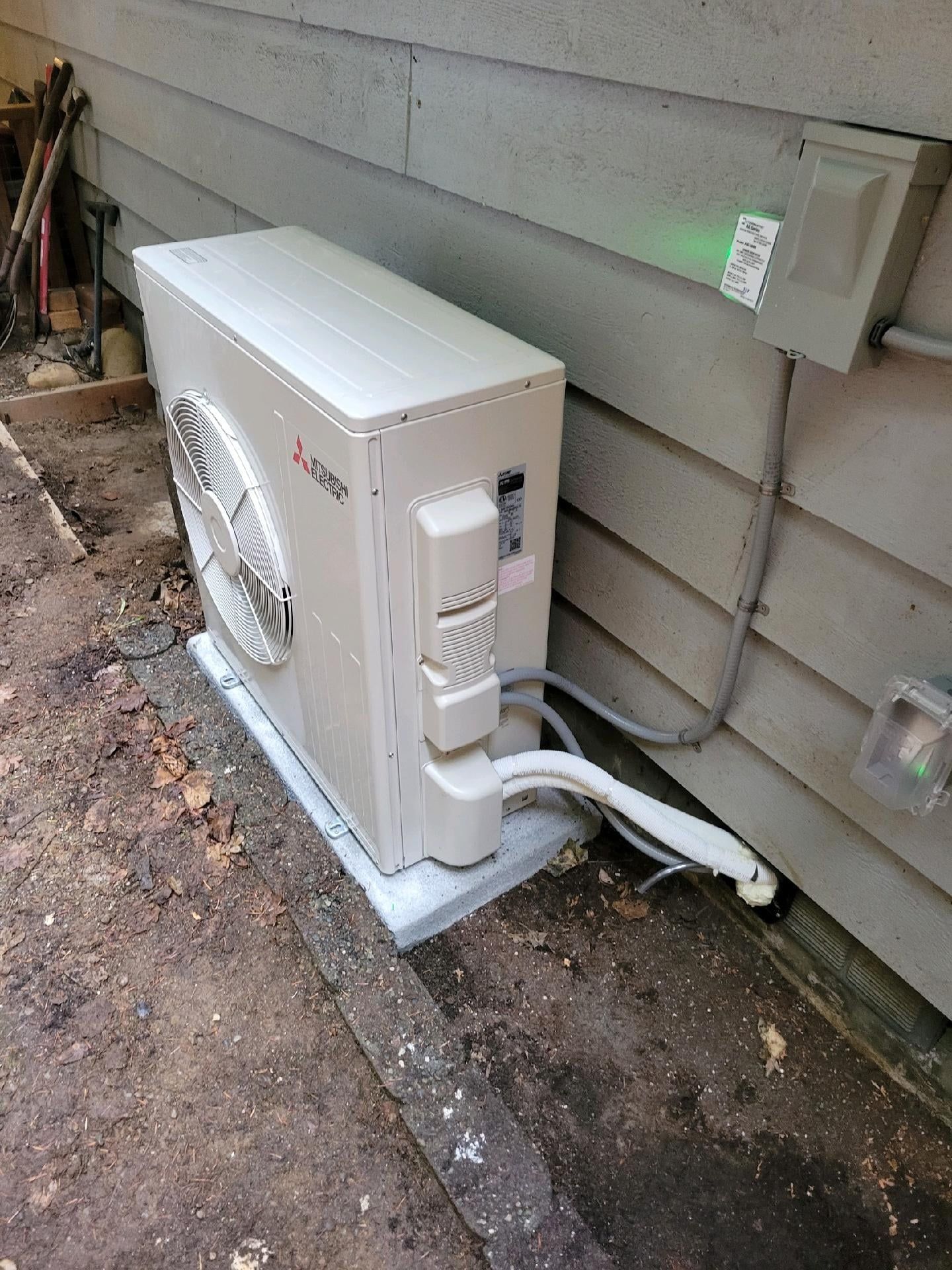 White HVAC unit mounted on concrete, next to a grey siding wall. Conduit and power box visible.